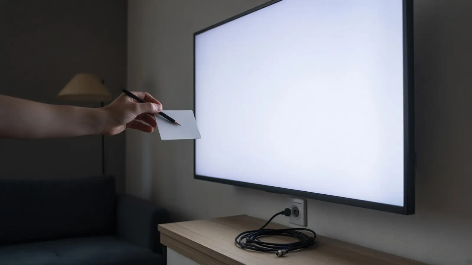 Eine Hand hält abends einen leeren Zettel und einen Stift nah an den hell leuchtenden Fernseher
